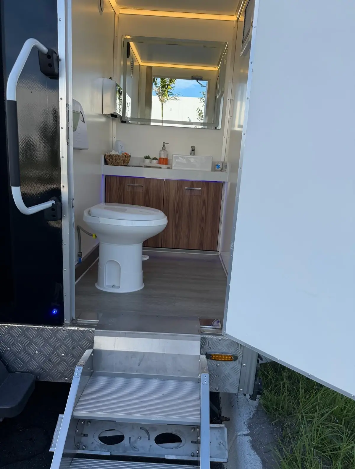 Open door of a restroom trailer station showing the toilet, sink and mirror